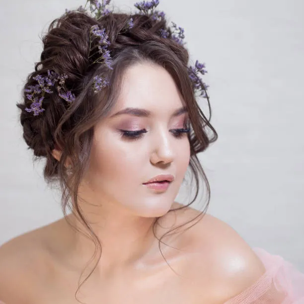 Three-quarter view of a bride wearing a delicate braided crown updo like a halo, long dark brown hair, lace wedding dress, outdoor ceremony with floral bokeh background.