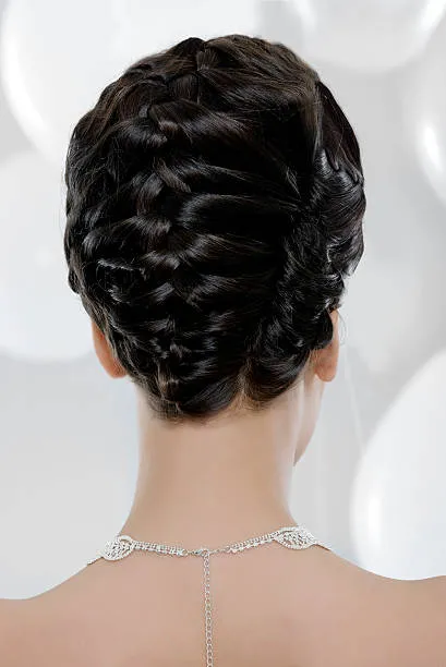 Back view of a bride with an elegant braided chignon, medium-length light brown hair, lace wedding dress visible, soft window light in elegant interior.