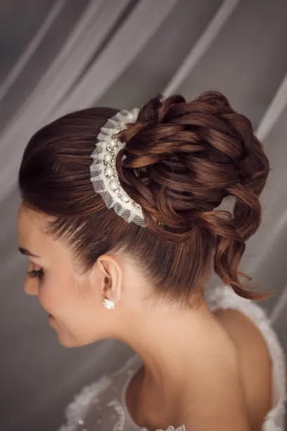 Side profile of a bride with an elegant high bun, statement earrings, dramatic neckline dress, soft studio lighting with neutral background.
