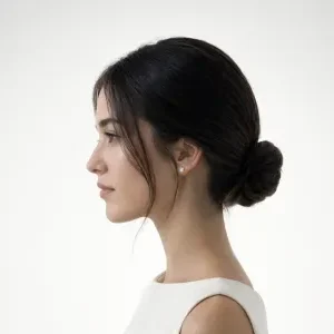 Side profile of a bride with a low ballerina bun, delicate face-framing pieces, straight dark hair, minimalist gown, clean white studio backgroun