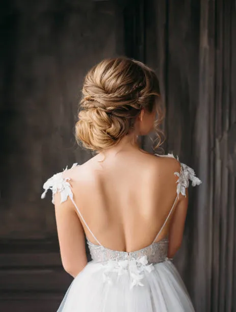 Back view of a bride with a twisted low bun, medium blonde hair with subtle texture, soft window light in an elegant interior with curtains.