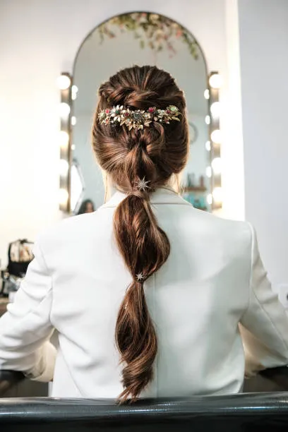 Back view of a bride with a braided halo bun, soft braid wrapping around crown flowing into low textured bun, lace gown, outdoor ceremony with soft natural light.