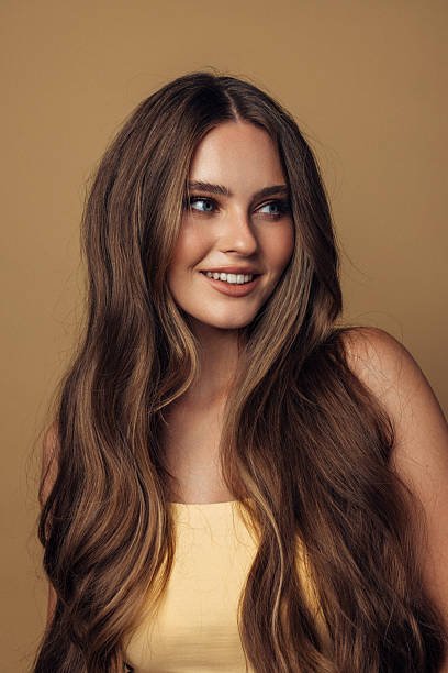 Portrait of a smiling woman with long brown hair wearing a yellow top in studio light Vibrant studio portrait featuring a smiling woman with long, flowing brown hair and blue eyes. She wears a soft yellow top against a warm beige backdrop, creating a friendly, approachable mood. Subtle makeup, natural glow, and confident posture convey beauty, fashion, and contemporary portrait style. hairstyles for female models stock pictures, royalty-free photos & images